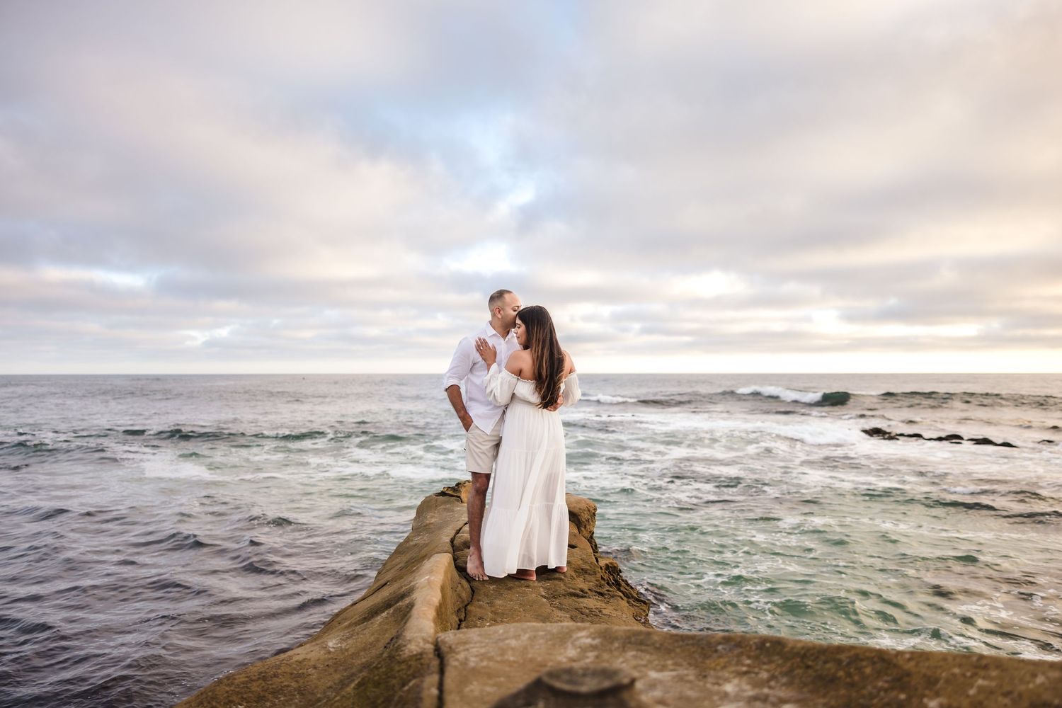 A couple shares a romantic moment on a rocky ocean jetty at sunset while waves crash around them.
