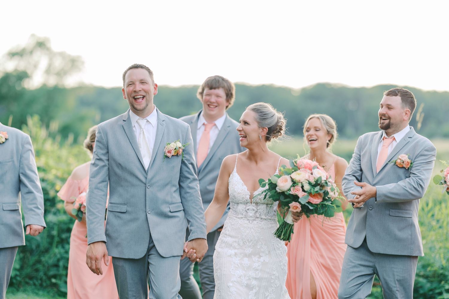 Wedding party in gray suits and peach bridesmaid dresses share a candid moment outdoors in summer.
