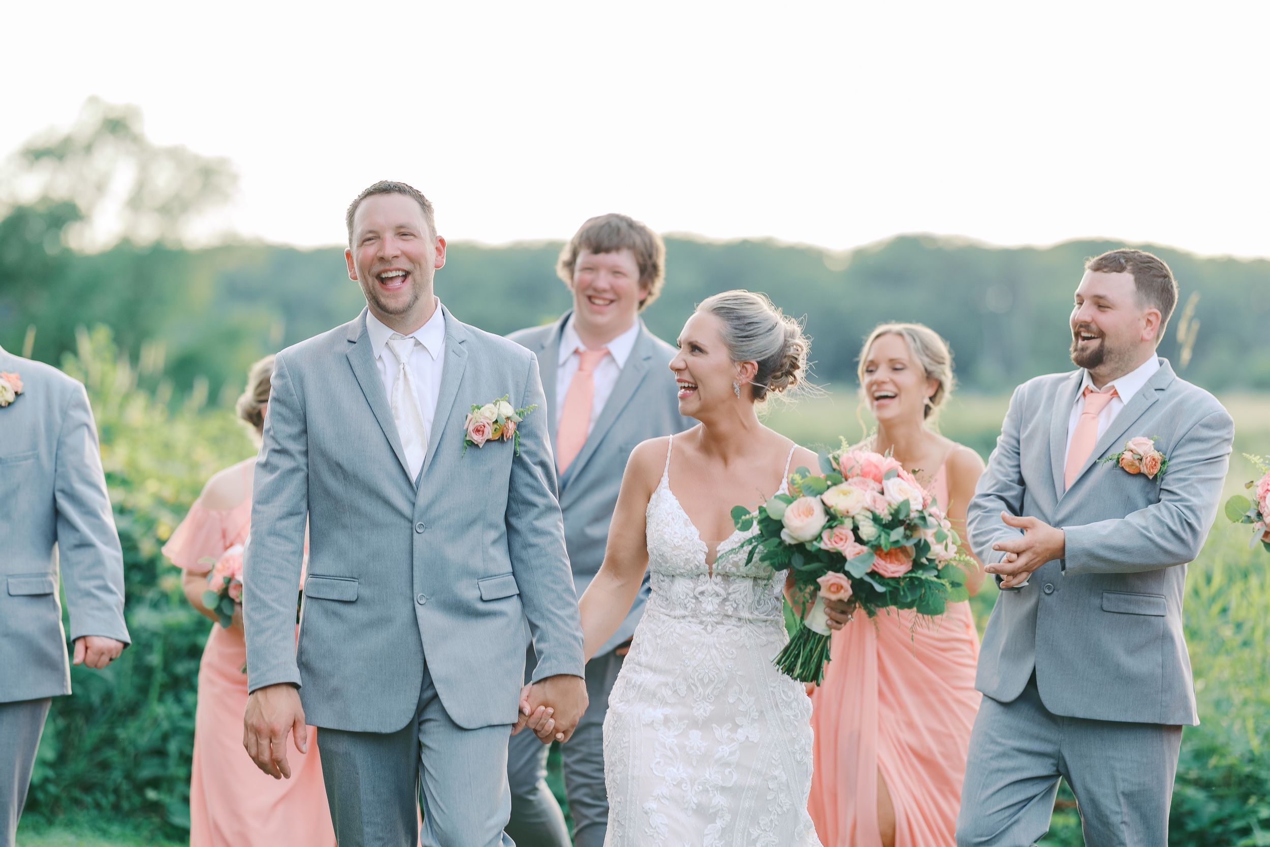 Wedding party in gray suits and peach bridesmaid dresses share a candid moment outdoors in summer.