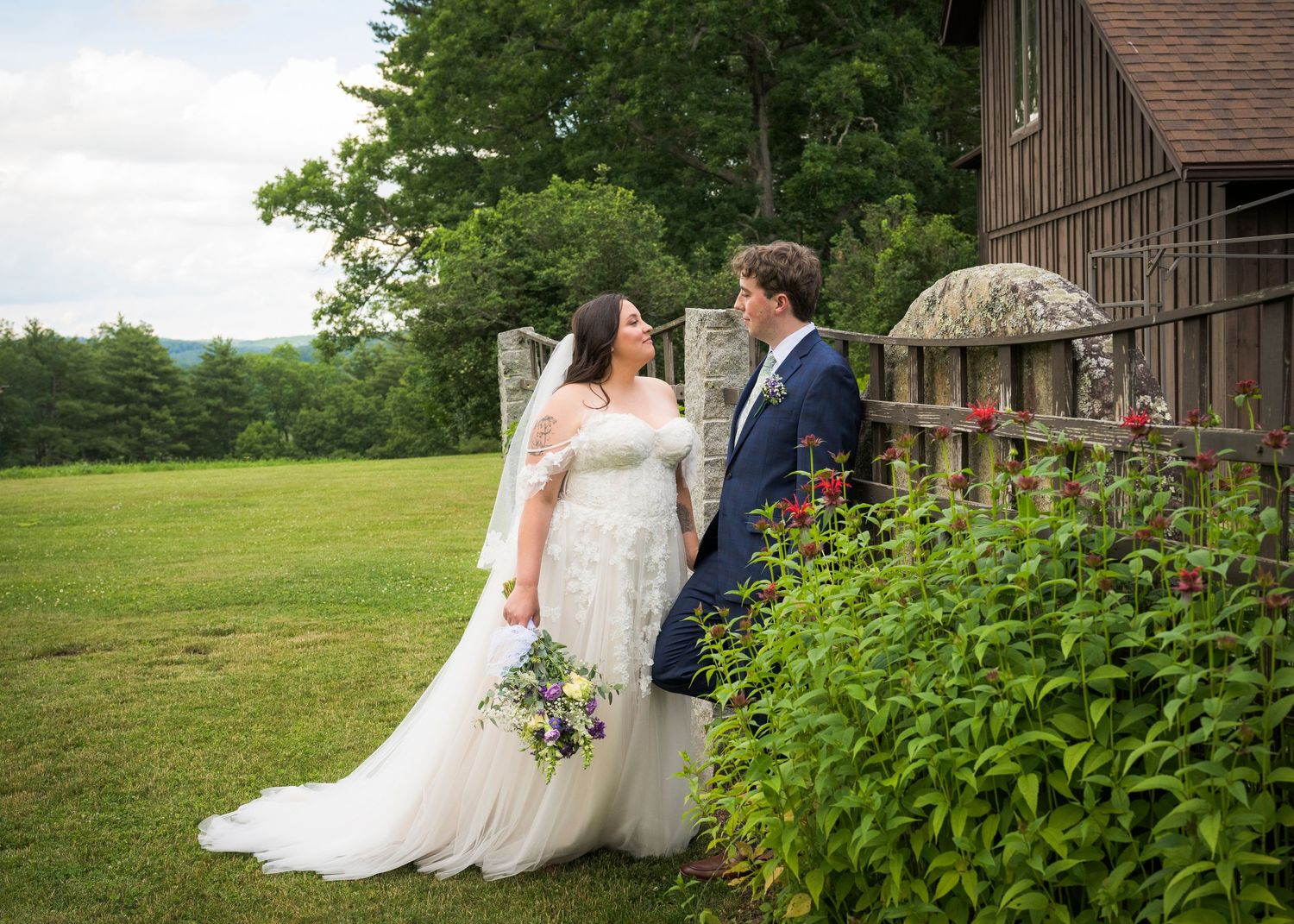 Newlyweds share an intimate moment outdoors with autumn foliage and rustic architecture in background.