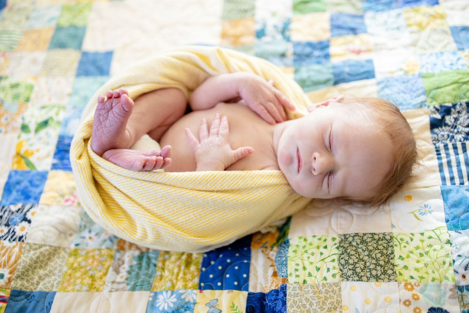 A sleeping newborn baby wrapped in a yellow blanket rests on a colorful patchwork quilt.