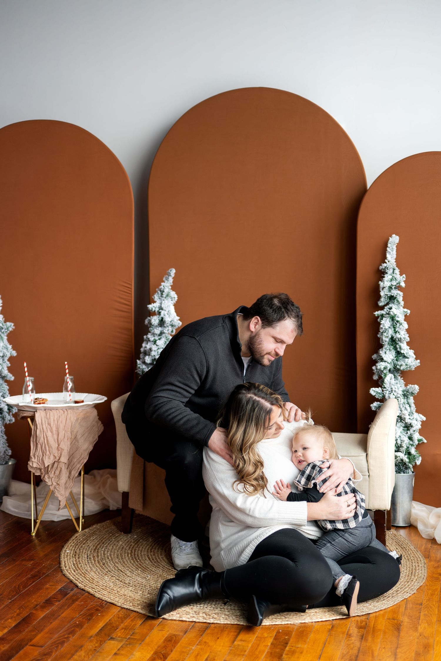 Father leans over mother and toddler son sitting on rug in front of brown arched Christmas backdrop.
