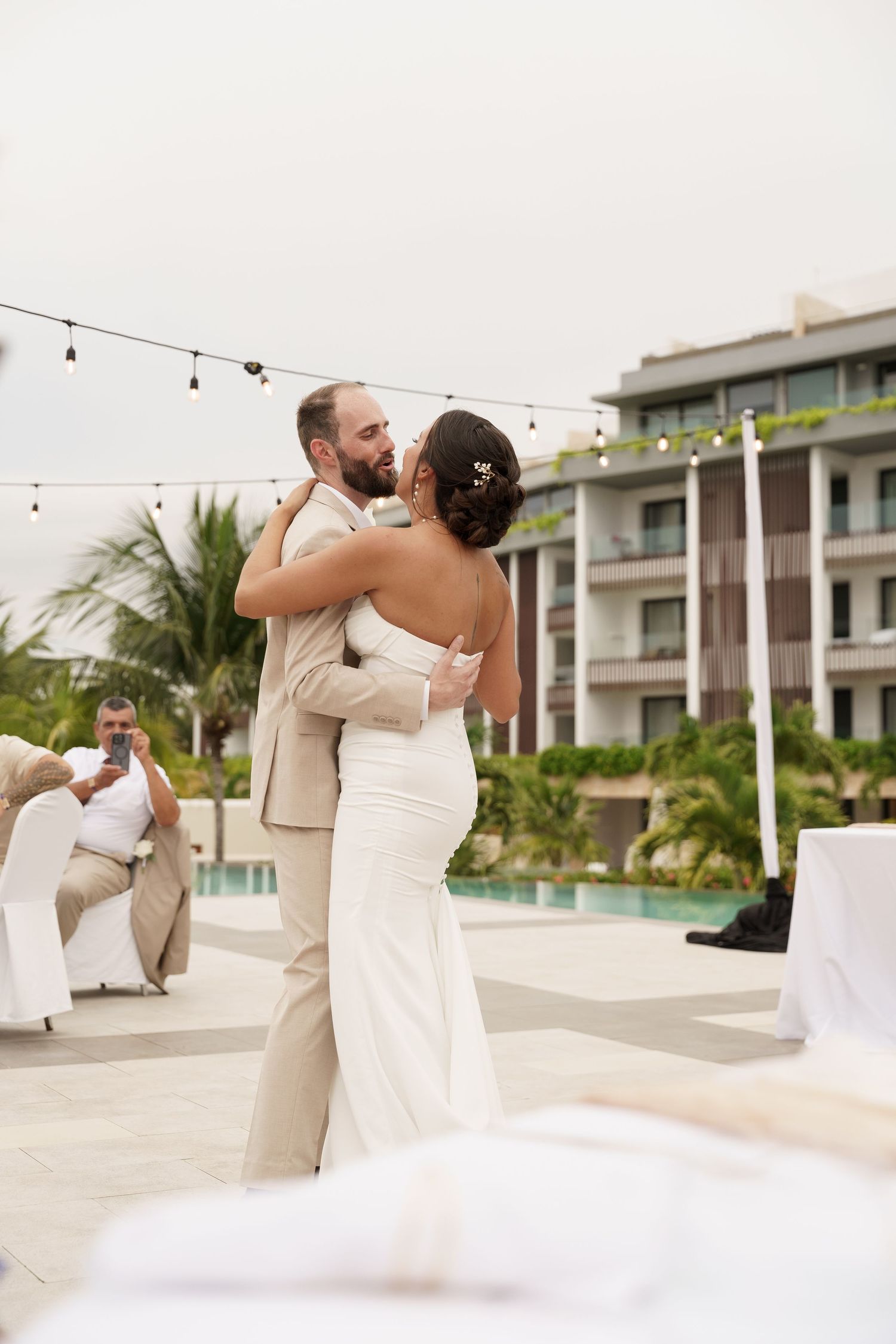 A couple shares an intimate dance moment on an outdoor terrace with modern buildings and palm trees in the background.
