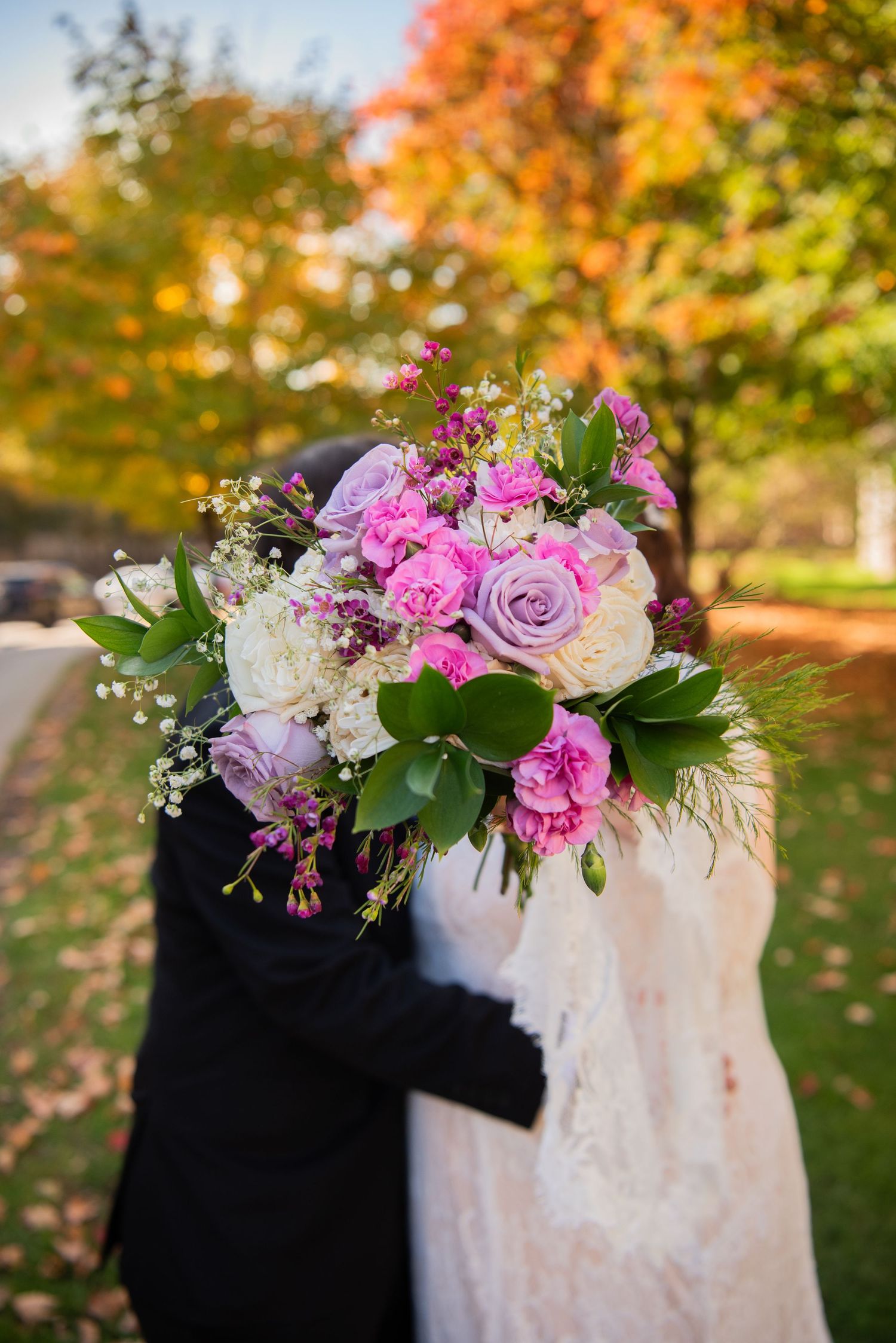 A beautiful wedding bouquet with pink and purple roses against a white dress and autumn foliage background.