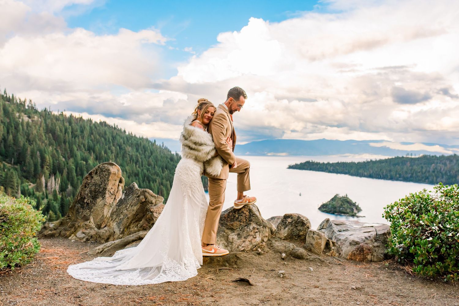 A couple in wedding attire embraces on a scenic mountain overlook with a lake and forest landscape in the background.
