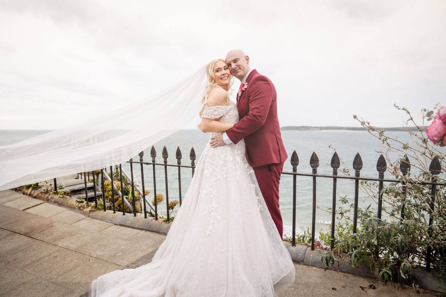 A romantic wedding moment captured outdoors along an iron fence with flowing white dress and burgundy suit.