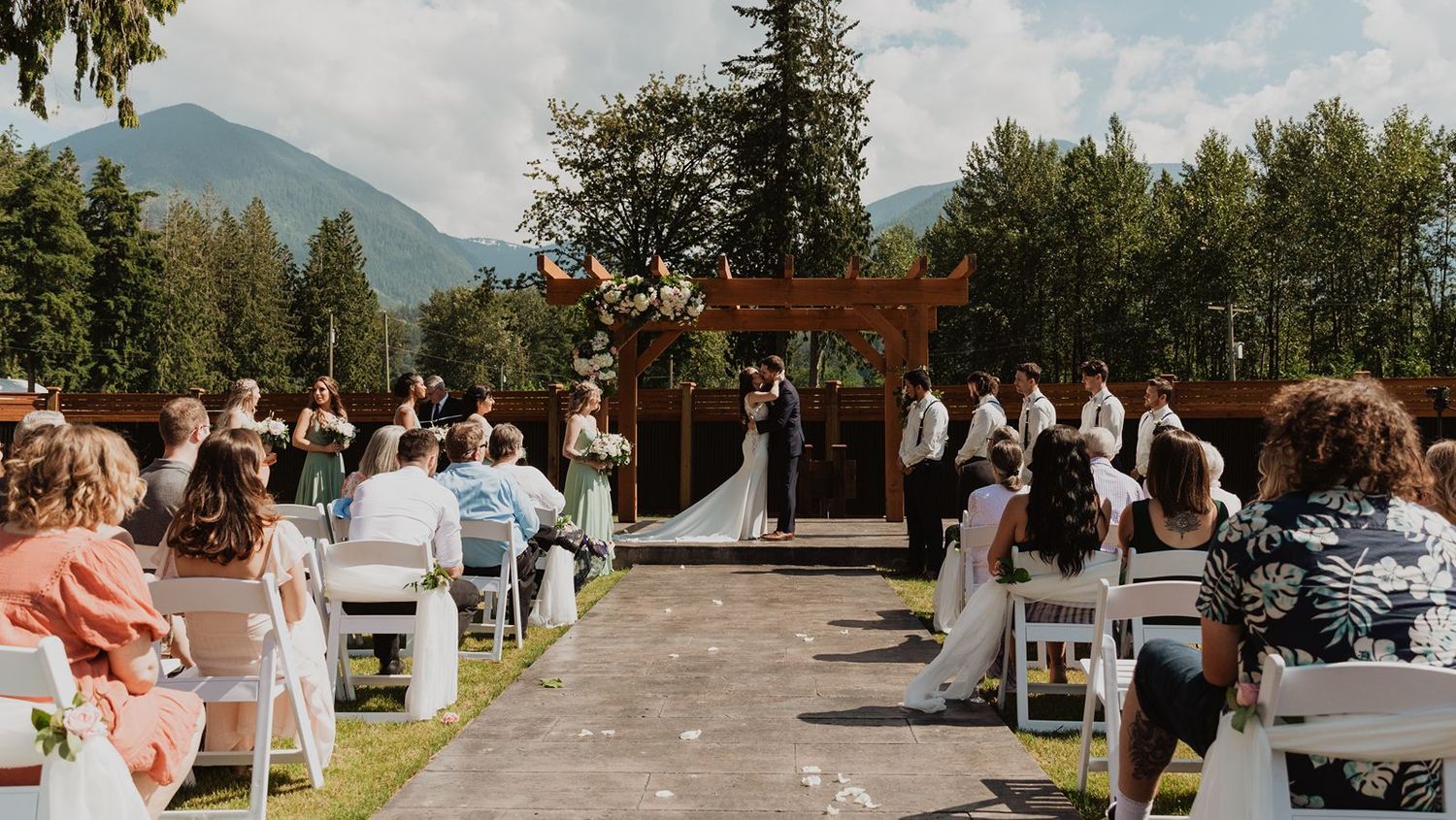 Outdoor mountain wedding ceremony with guests seated on white chairs and scenic forest backdrop.