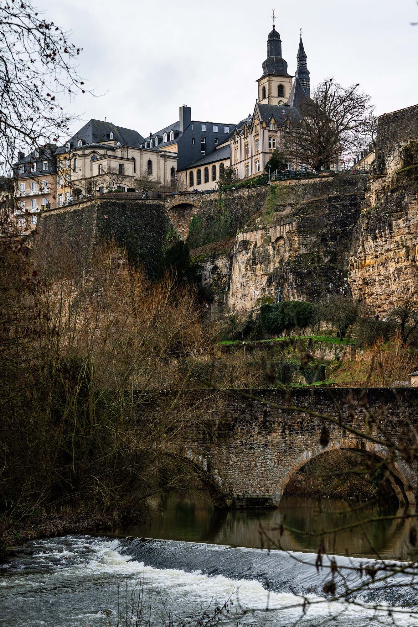 Historic stone walls and buildings of Luxembourg City perch above the Alzette River with a small waterfall in the foreground.
