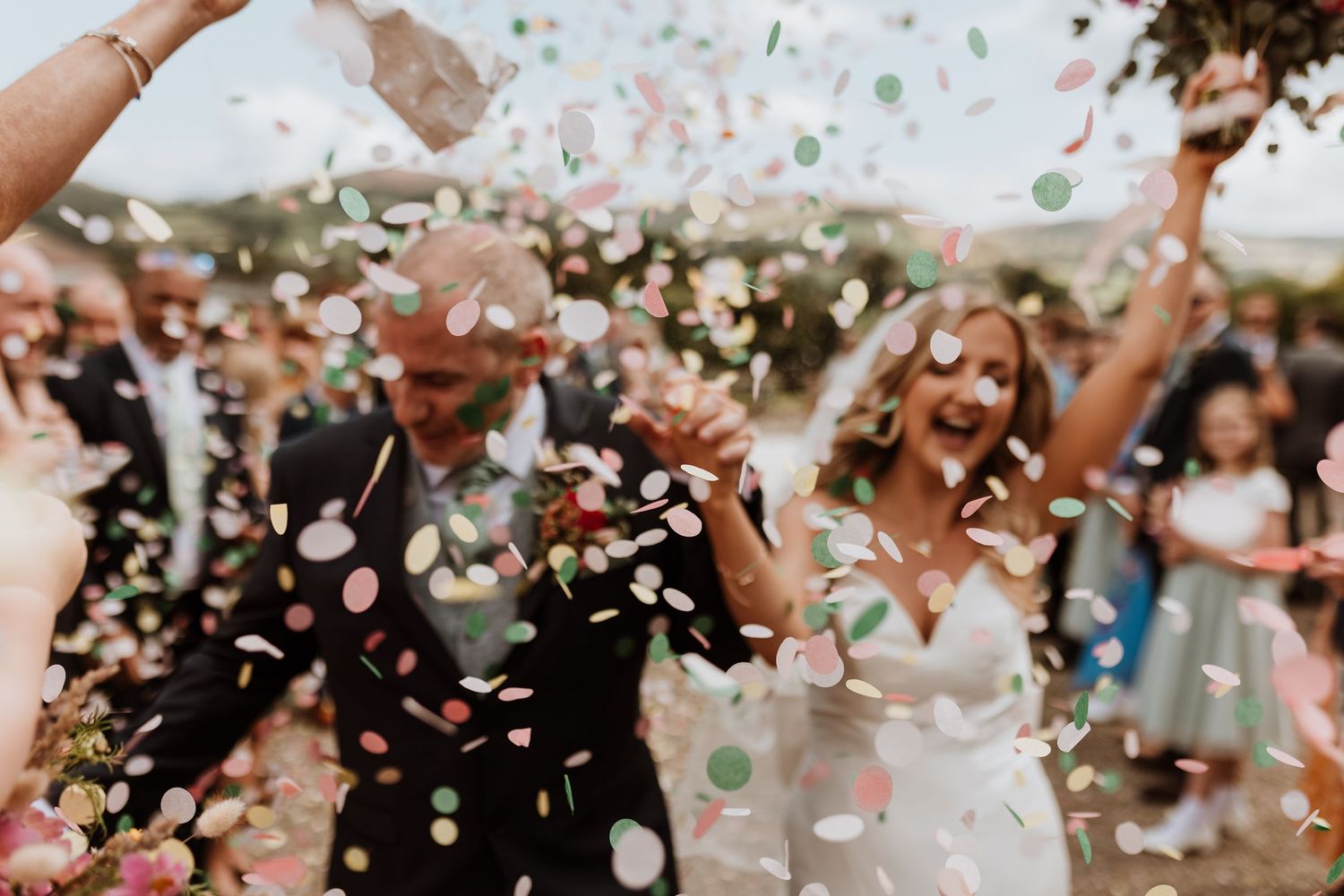 Newlywed couple celebrates under a shower of colorful confetti surrounded by cheering guests at an outdoor wedding.