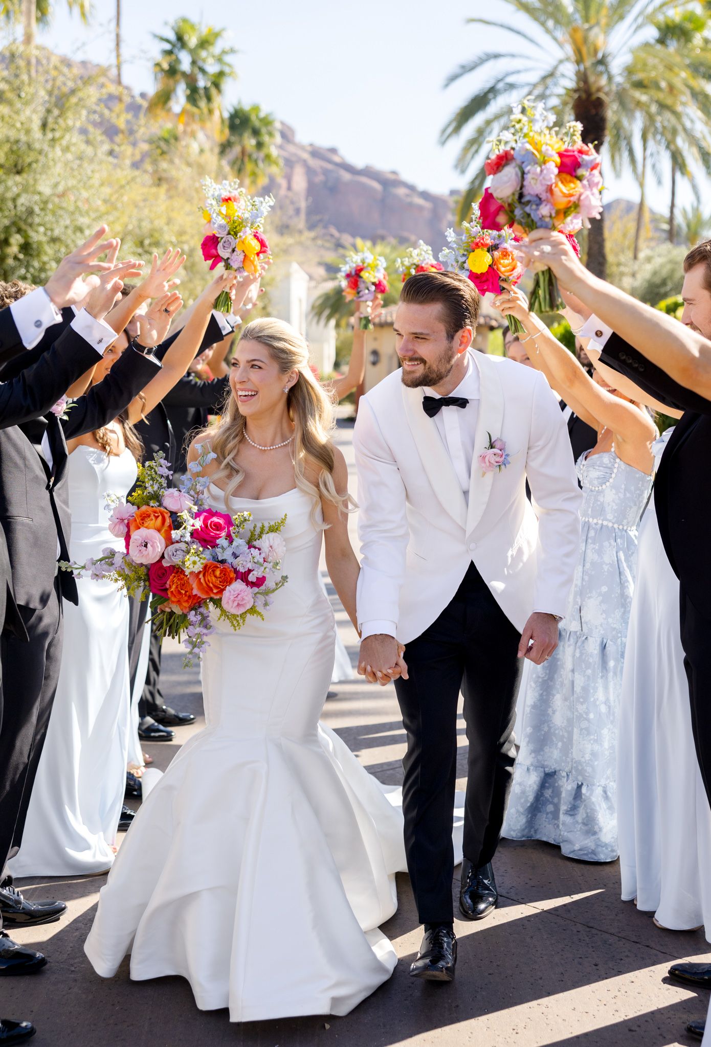 Wedding party celebrates with colorful bouquets and raised hands while walking together under palm trees on a sunny day.