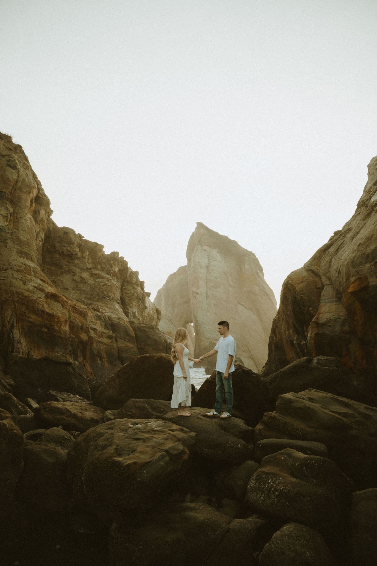 Two figures stand silhouetted against dramatic rocky cliffs during golden hour creating a moody coastal landscape scene.