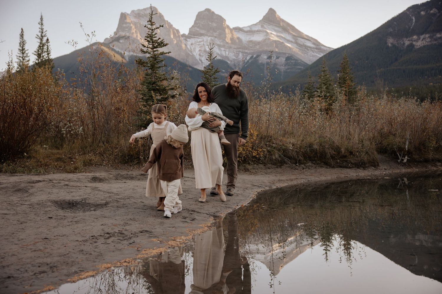 A couple stands near a reflective mountain lake with Three Sisters peaks towering in the background at sunset.