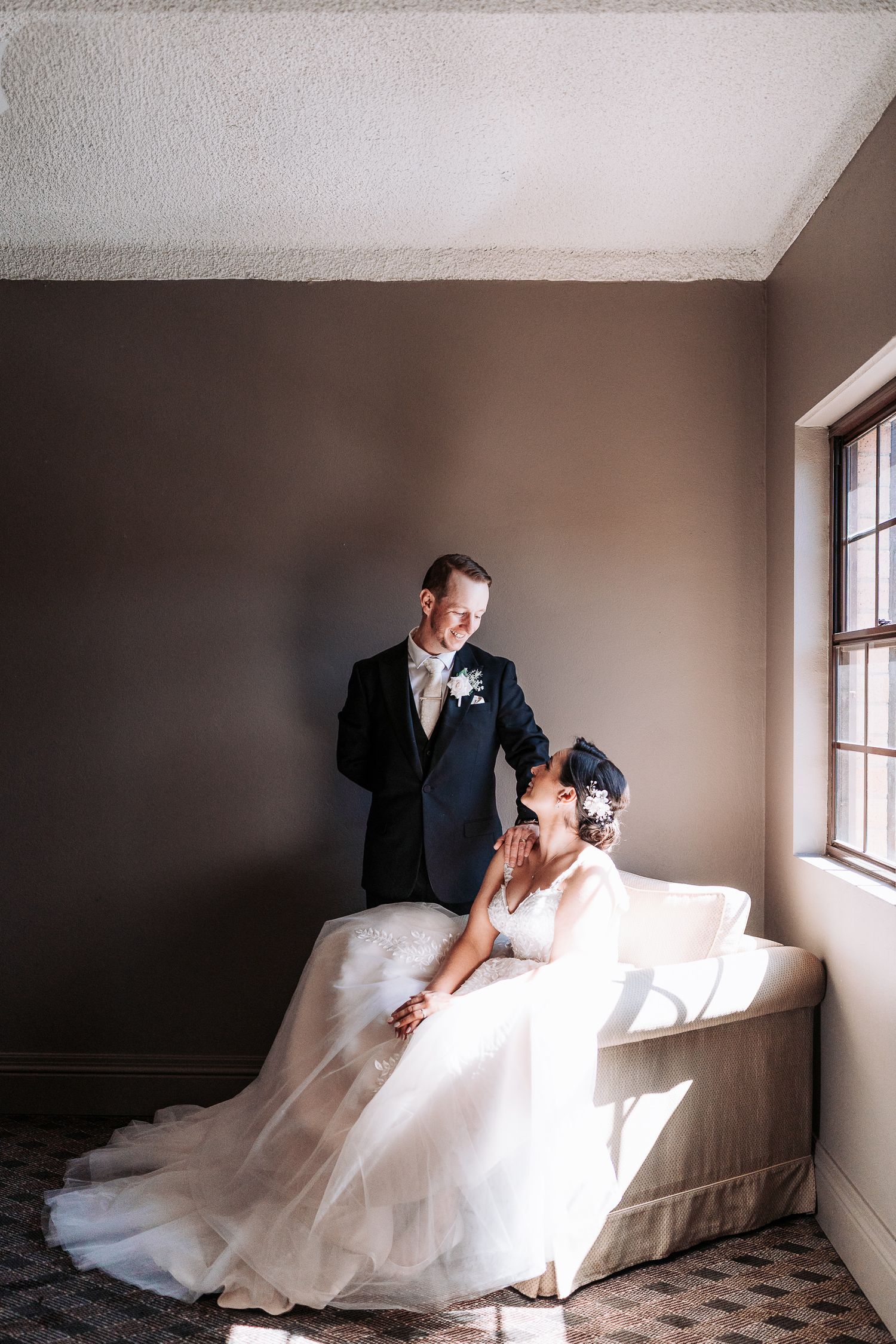 A romantic wedding couple lounges together on a white couch in a minimalist room with natural window lighting.