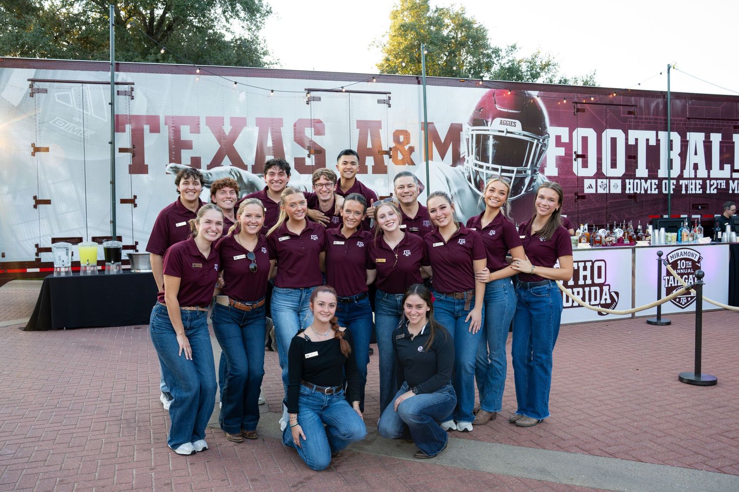 A group in maroon shirts poses together in front of a Texas A&M Football trailer display.
