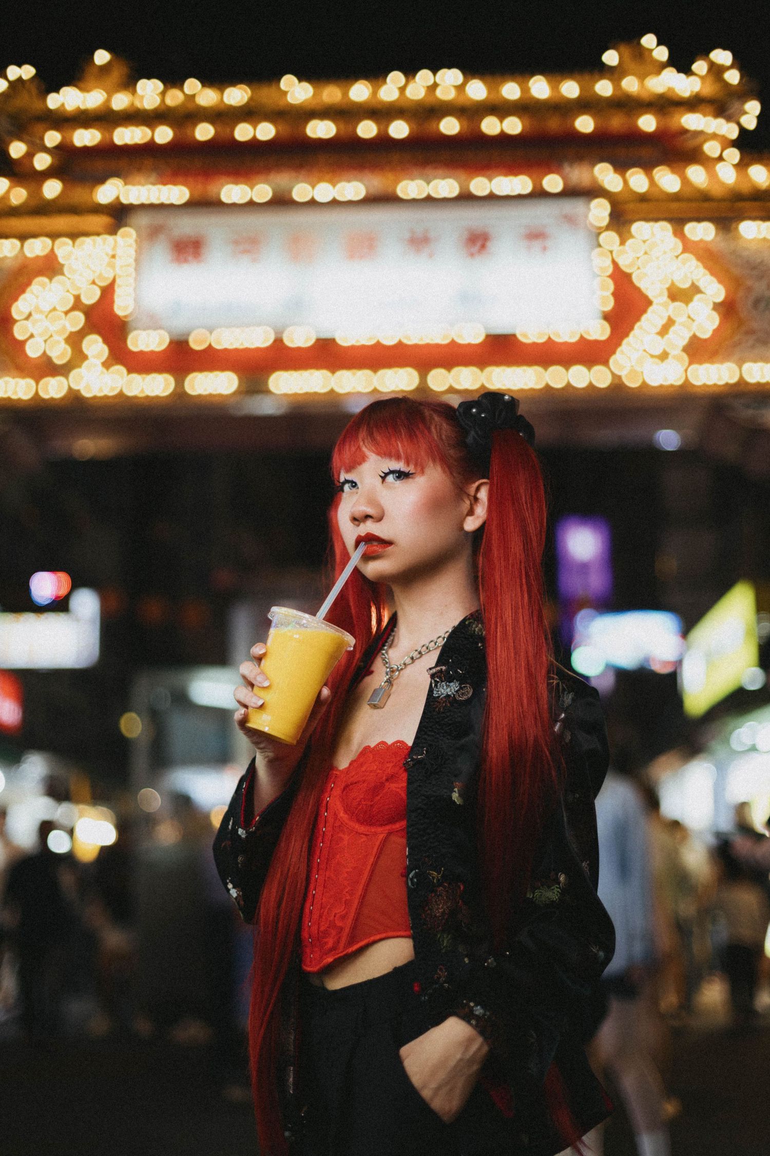 Young woman with red hair sipping orange juice under bright neon signs at a vibrant night market.