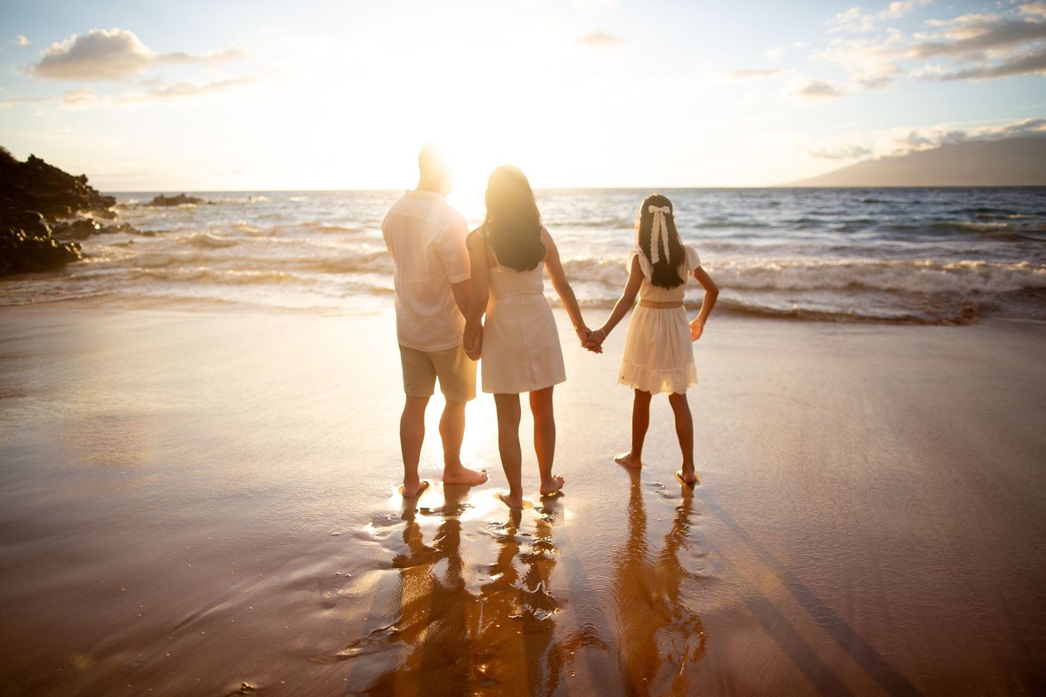 Family of three holding hands on a sandy beach at golden sunset with ocean waves in the background.