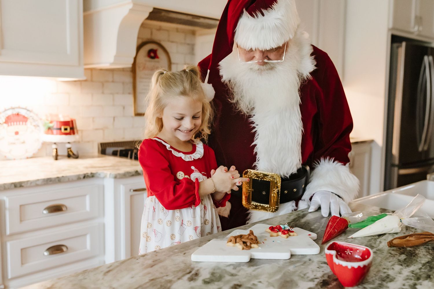A child and Santa Claus bake Christmas cookies together in a bright white modern kitchen.
