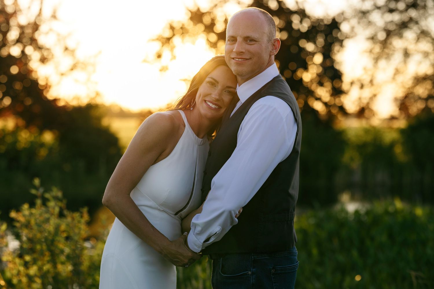 A couple embraces in golden sunset light during their wedding photoshoot in a meadow with trees in the background.
