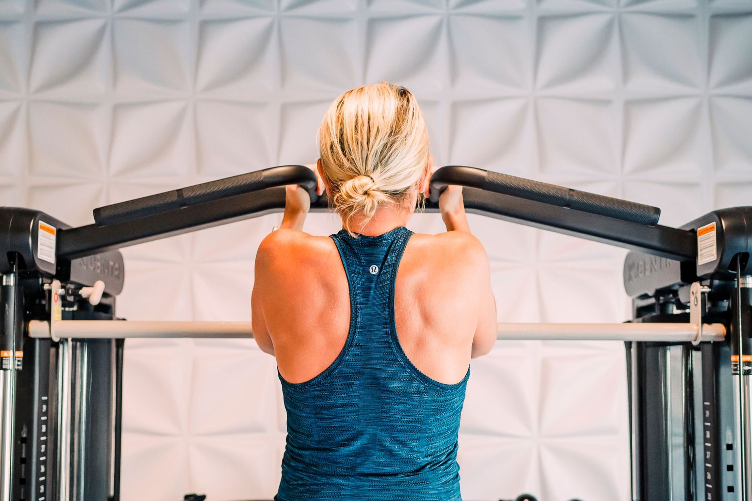 Person in blue tank top performing lat pulldown exercise at gym with geometric white wall pattern in background.