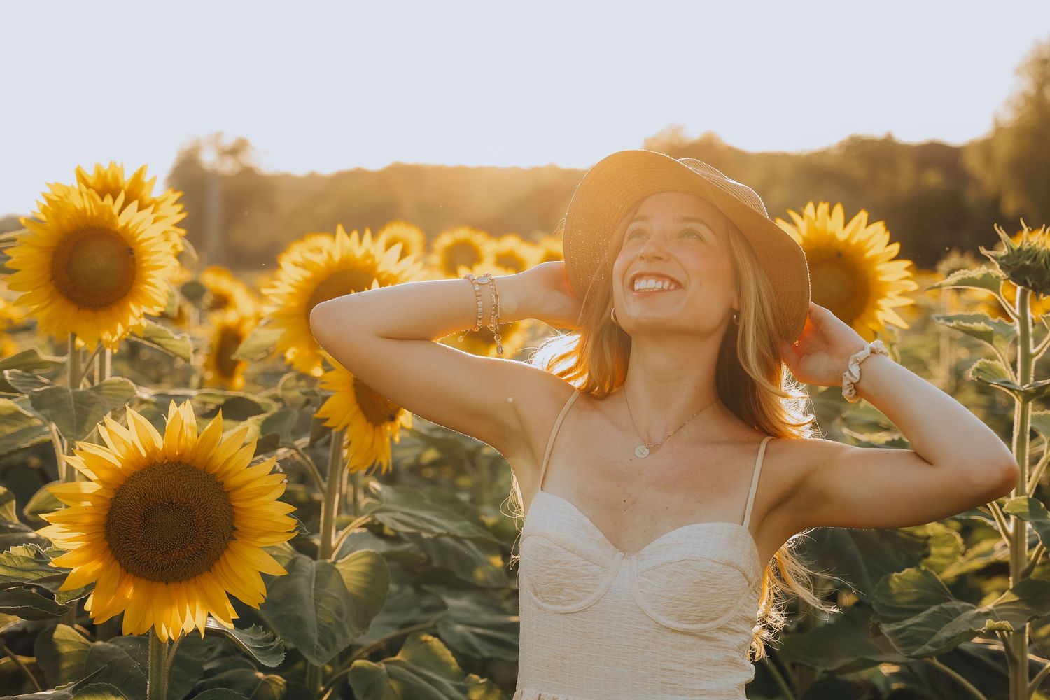 Happy woman in hat and white dress laughing in sunflower field at golden hour.