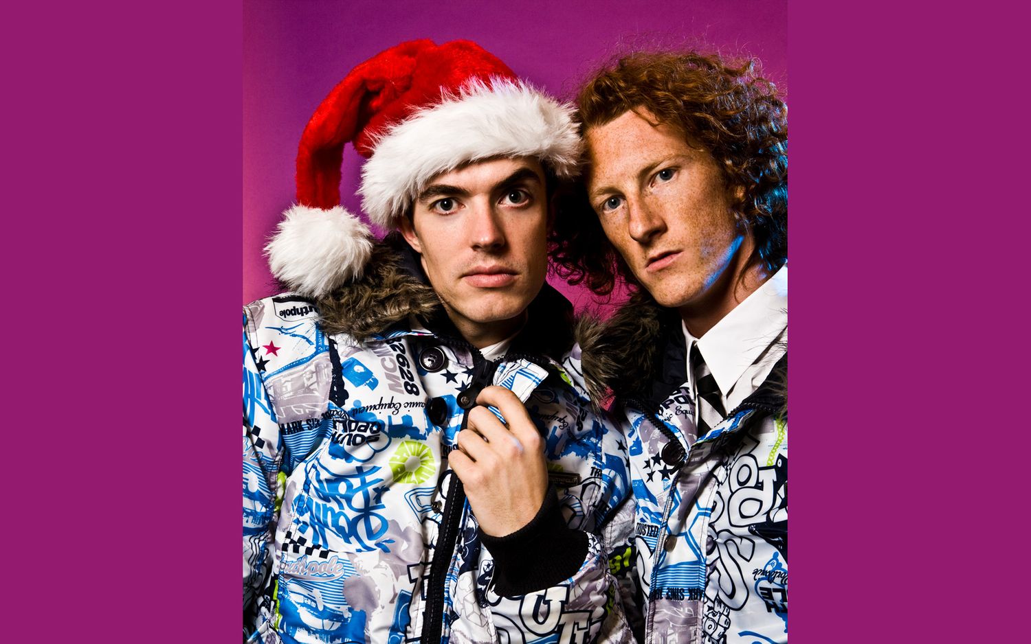 Two people wearing festive Santa hats and winter jackets with blue and white graffiti-style patterns pose together.