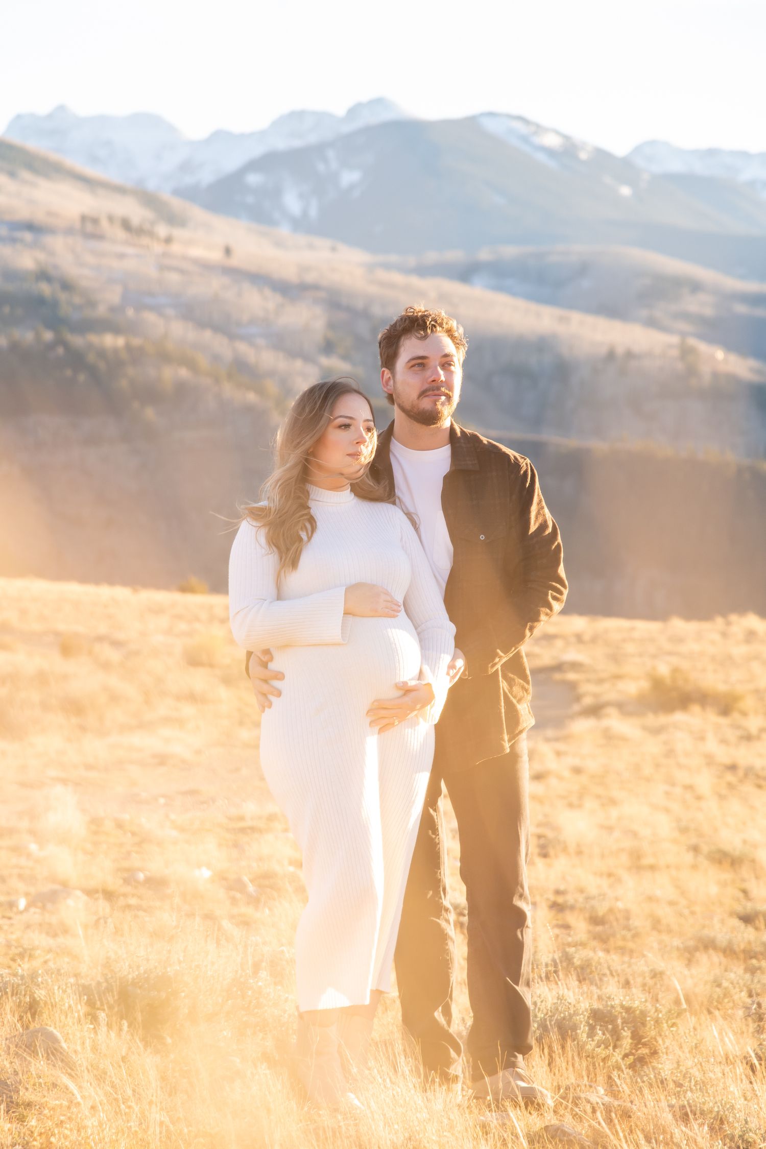 A couple stands together in a golden mountain meadow at sunset with mountains in the background.