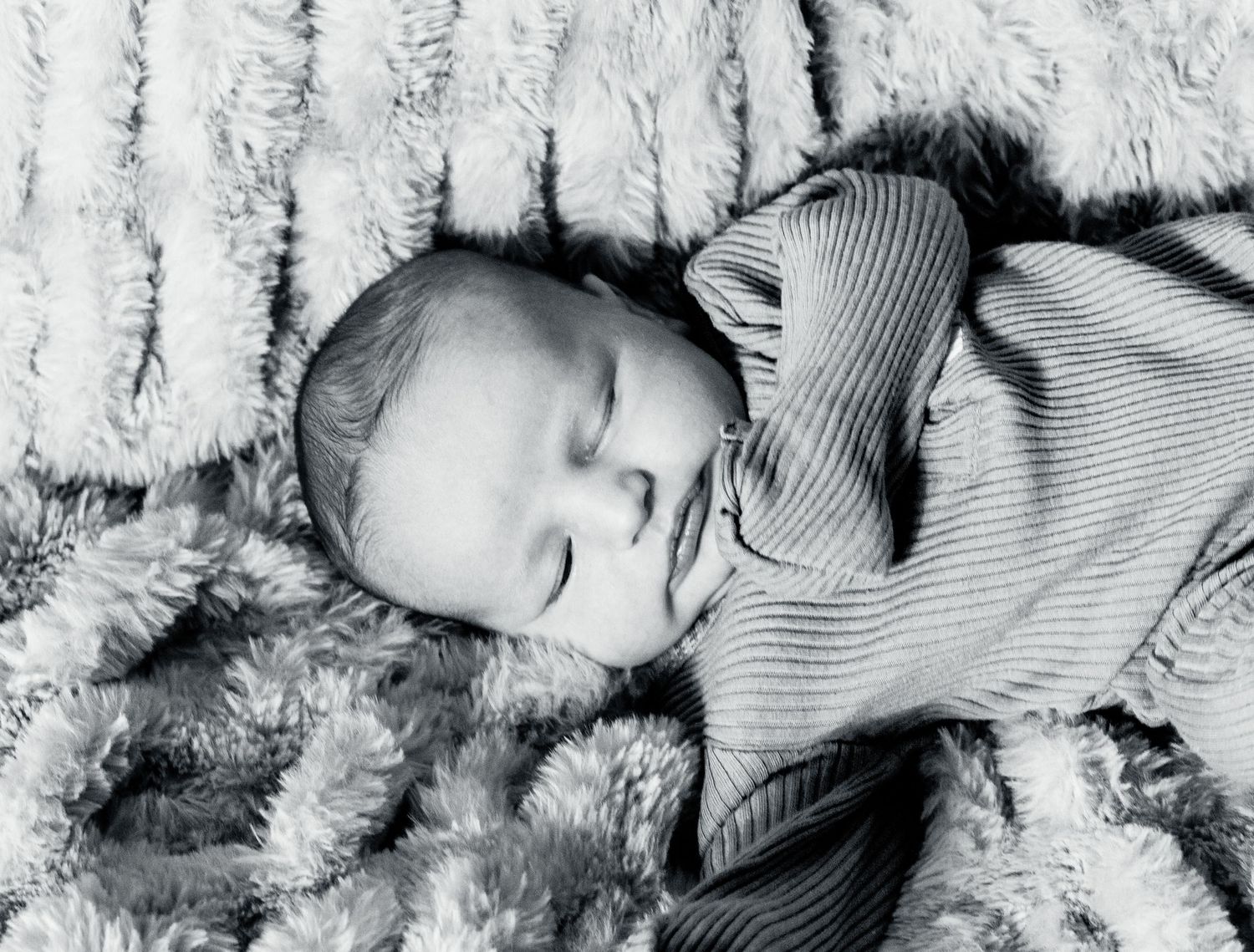 A peaceful sleeping baby wearing a striped outfit lies on a soft furry blanket in black and white.