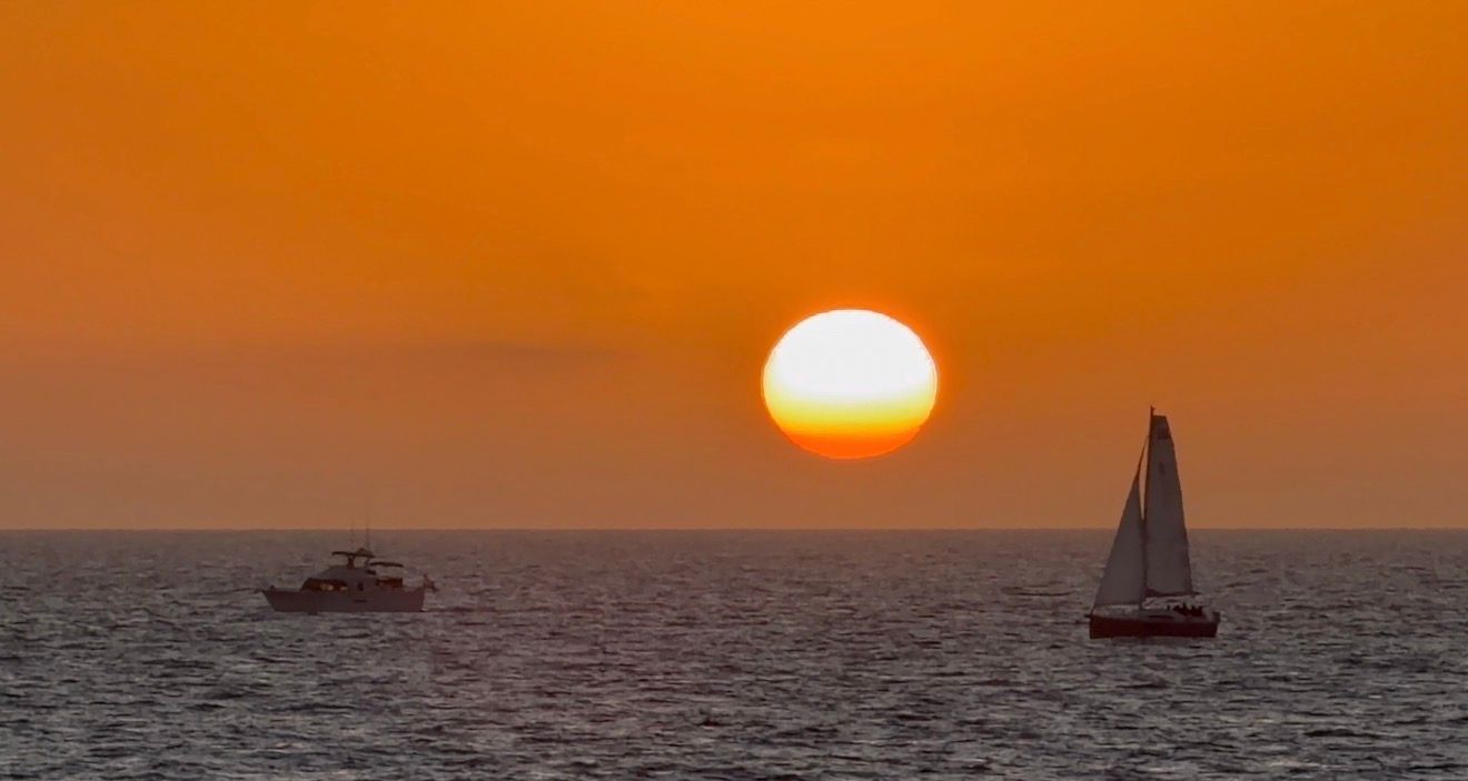 A vibrant orange and yellow sunset illuminates the horizon over calm ocean waters with silhouetted boats in the distance.