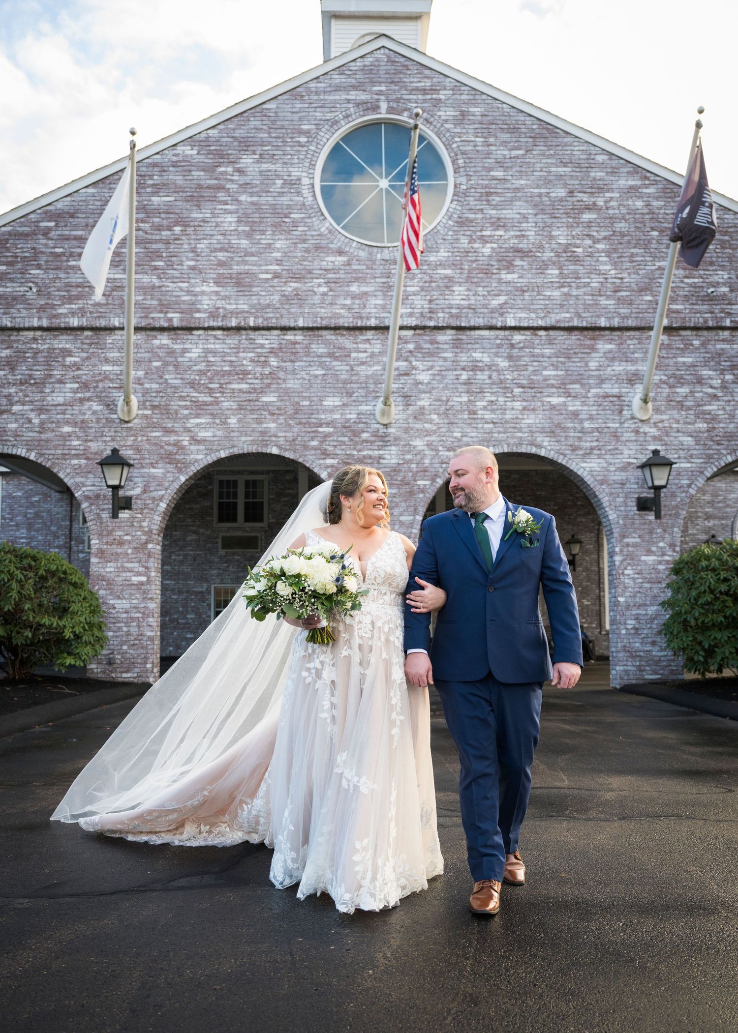 A wedding couple walks together outside a stone church building with arched entrances and flags flying above.