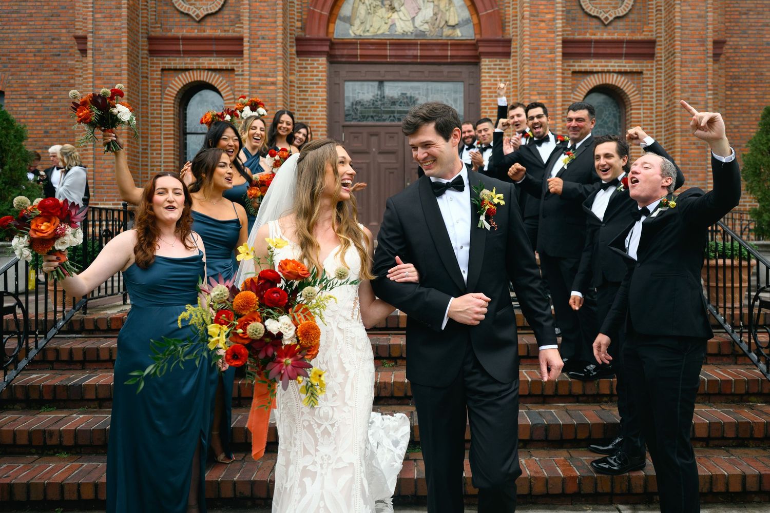 Wedding party in formal attire laughs together outside historic brick building with colorful autumn flowers.