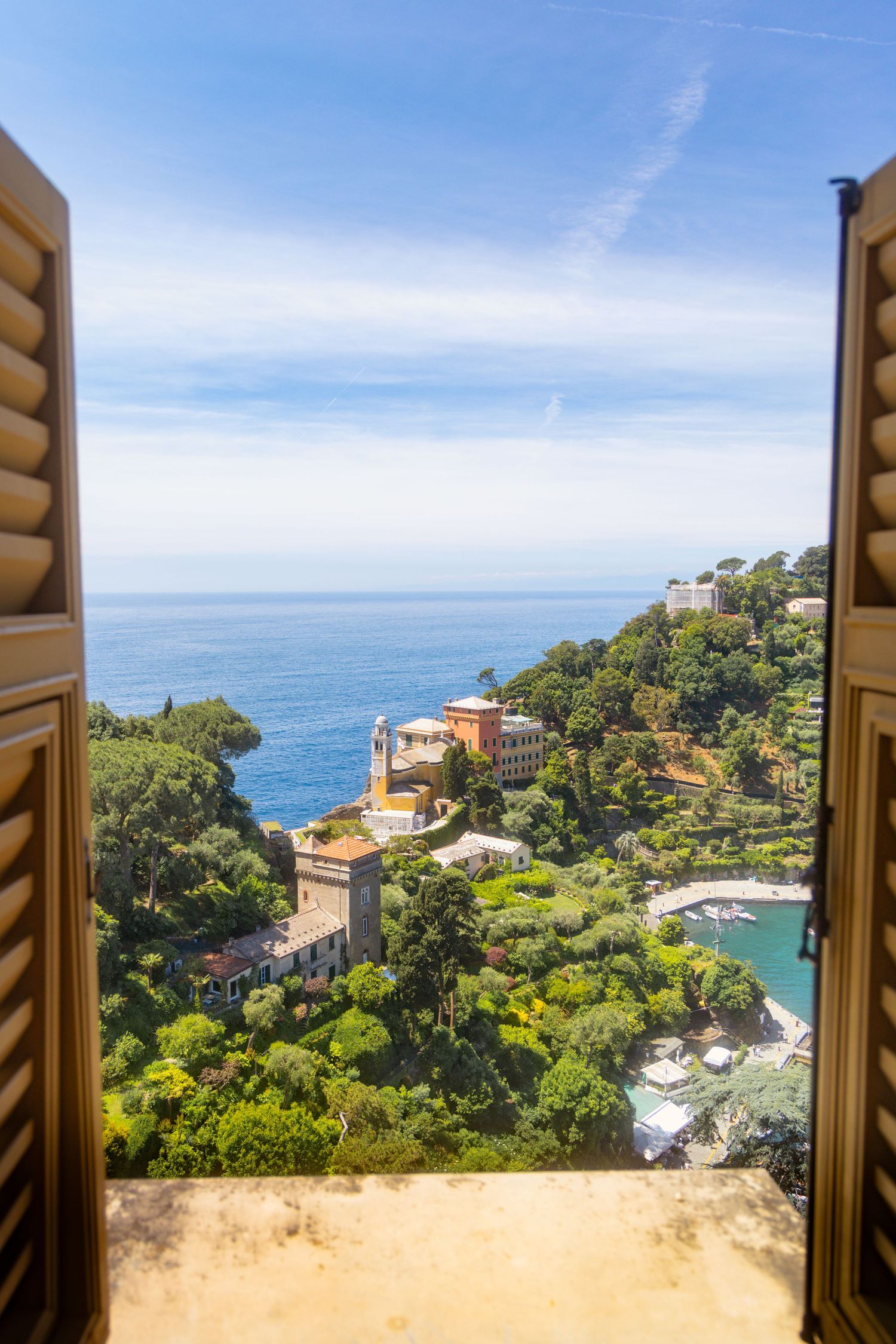 Mediterranean coastal view through open wooden shutters overlooking blue waters and villas.