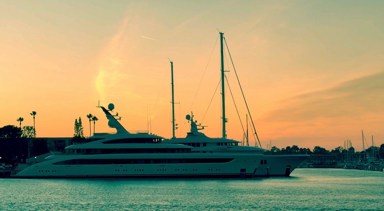 A panoramic view of a harbor at sunset with silhouettes of boats and cranes against an orange and teal sky.