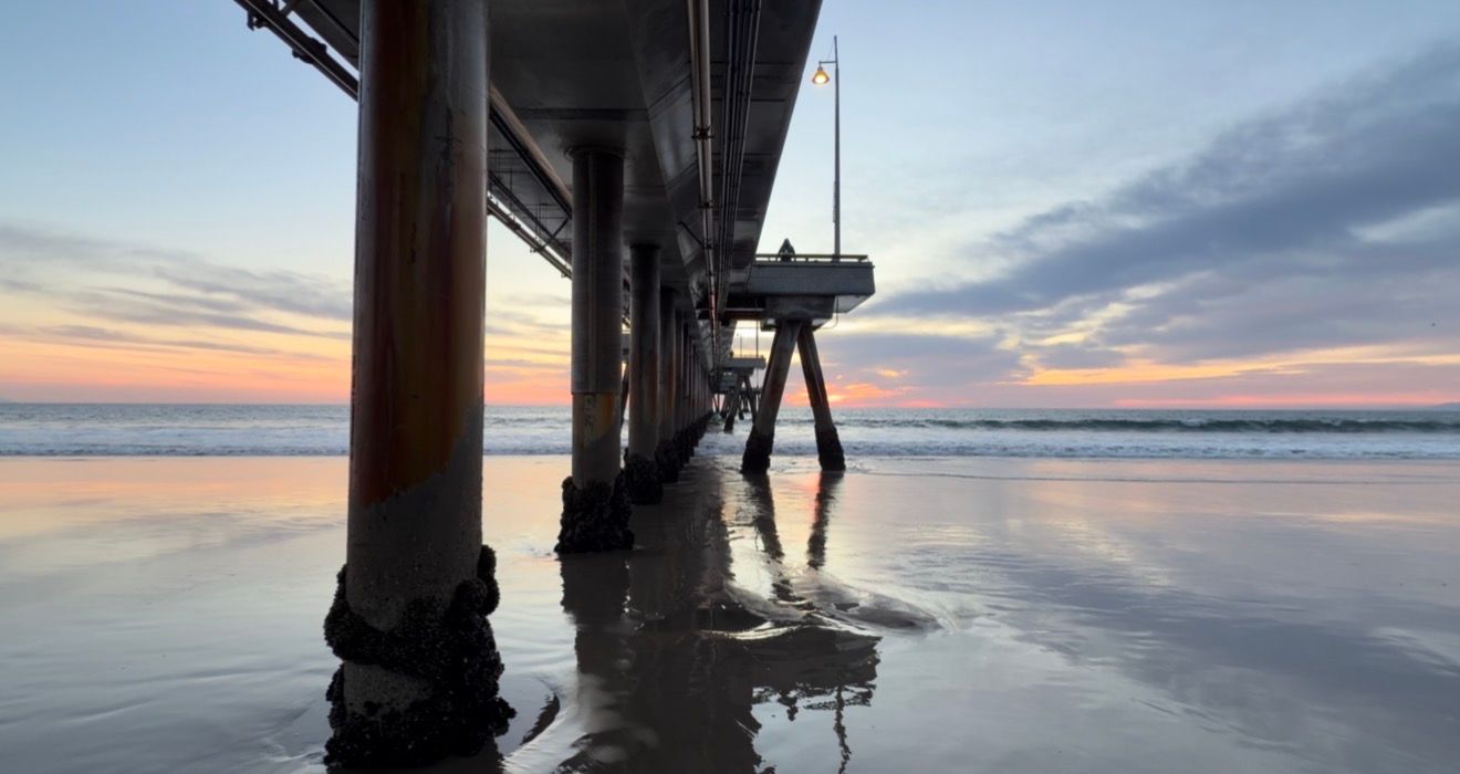 Silhouetted pier pilings reflect in calm ocean waters during a vibrant sunset with colorful clouds on the horizon.