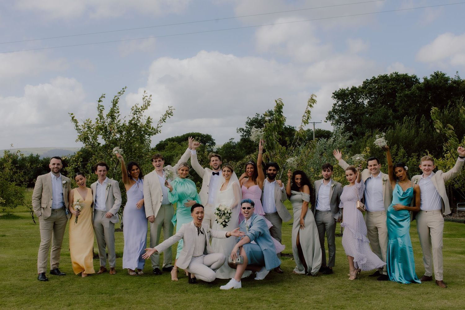 Wedding party in coordinated pastel outfits poses together on a grassy lawn under cloudy skies.