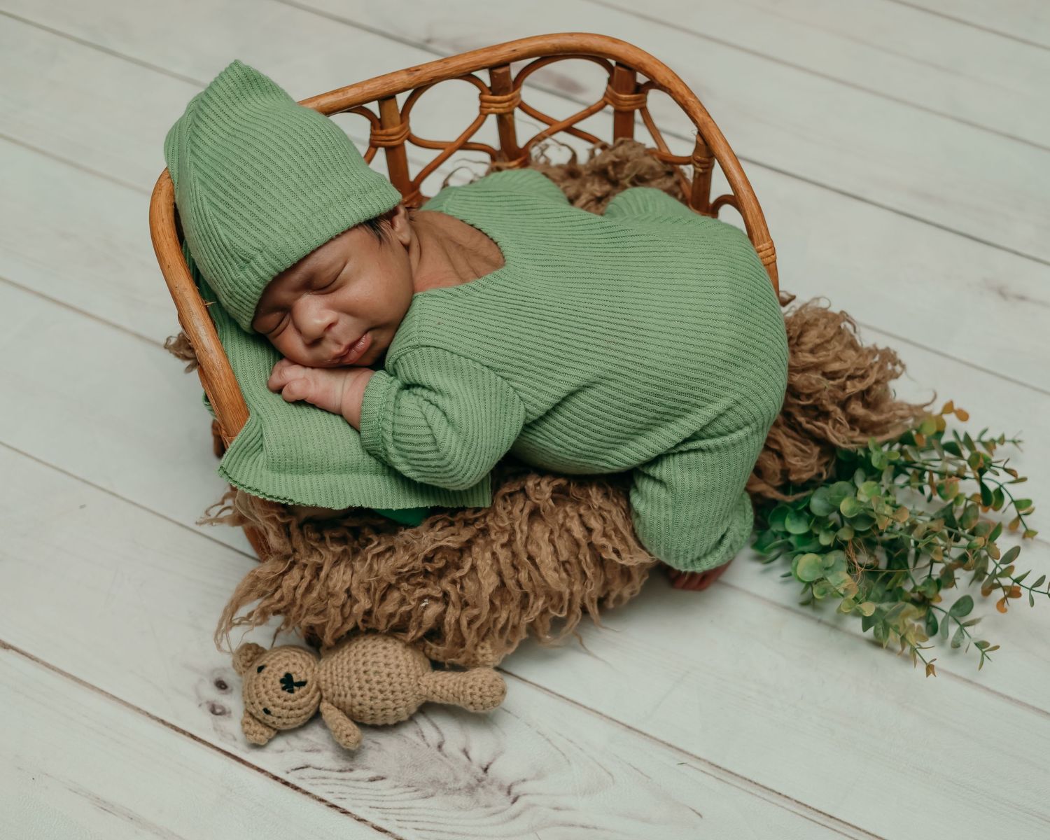 A sleeping baby in green knitwear curled up in a wicker chair with brown textured blanket.