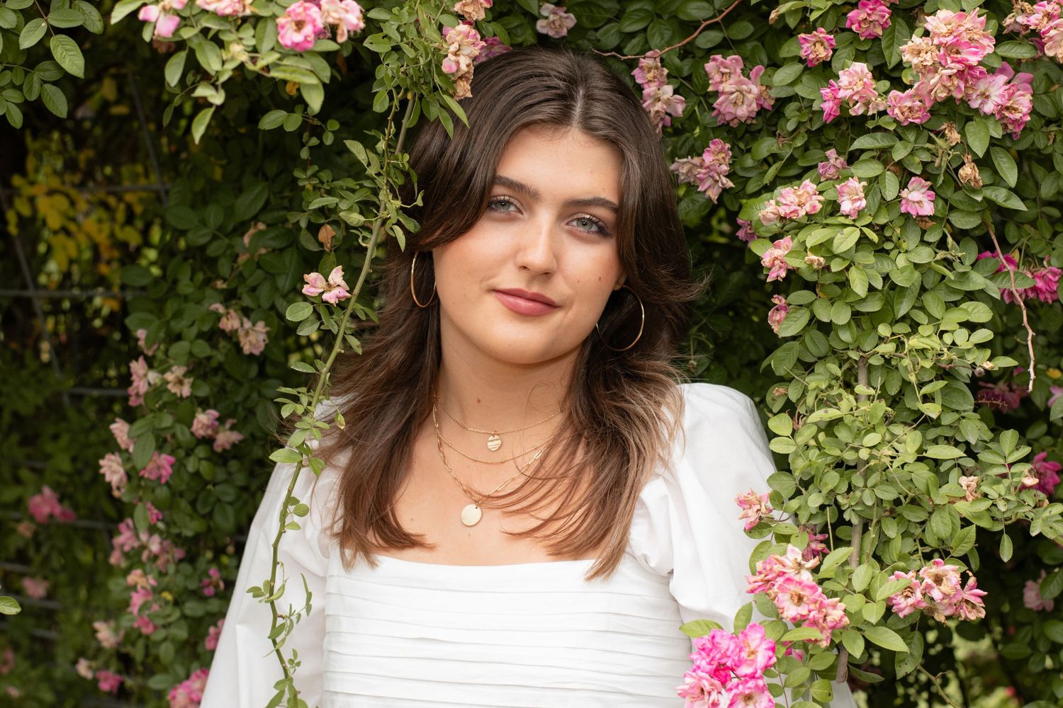 A person in a white dress poses among blooming pink roses in a garden.