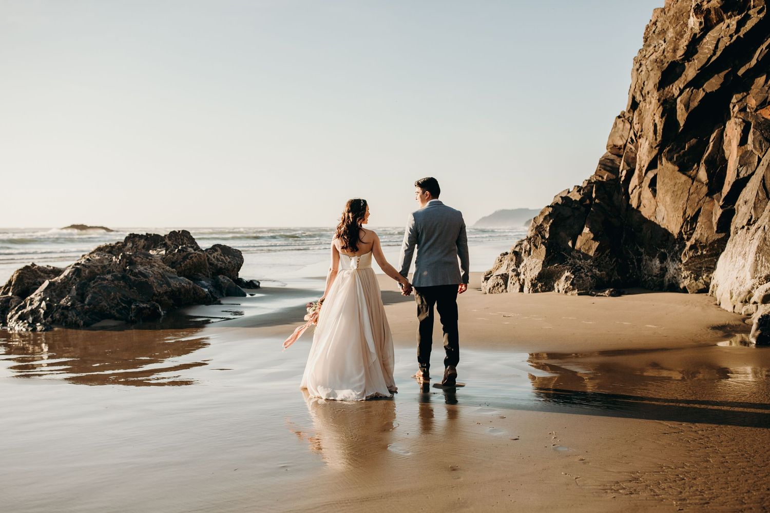A couple walks hand in hand along a scenic beach with rocky cliffs.