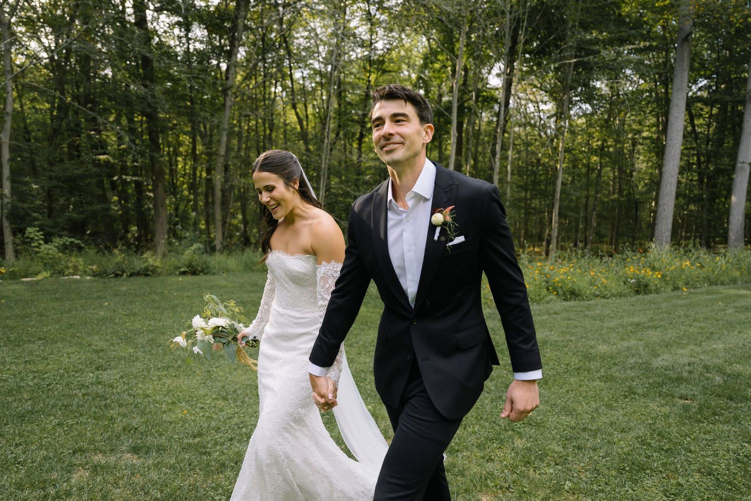 Couple walking hand in hand through a green meadow in wedding attire.