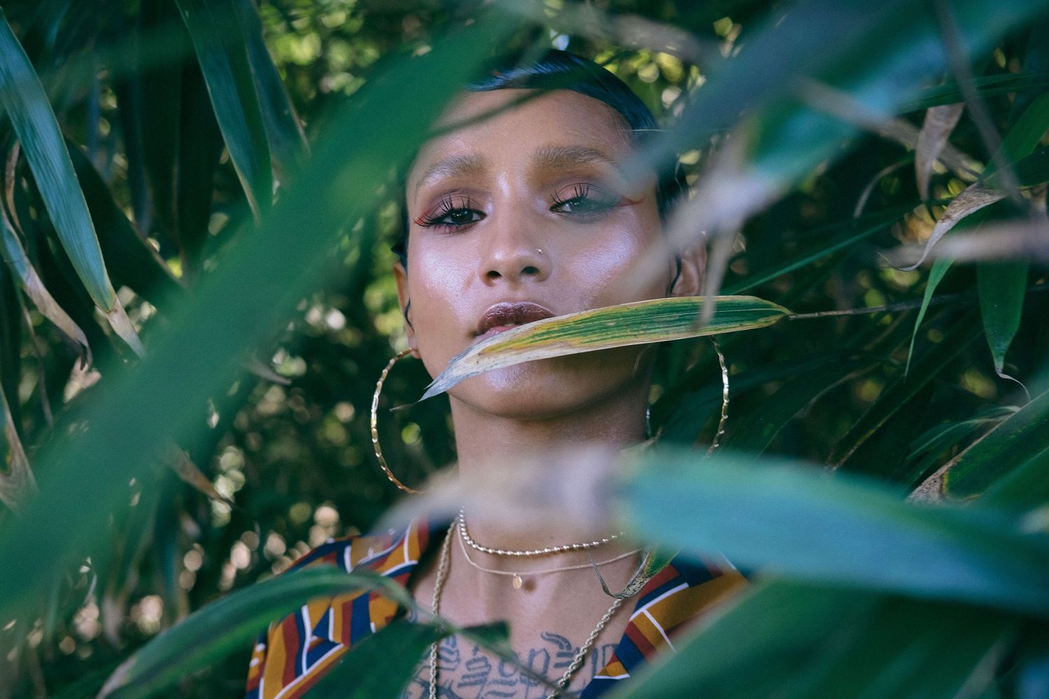 Artistic portrait through green leaves showing glittery makeup and jewelry details.
