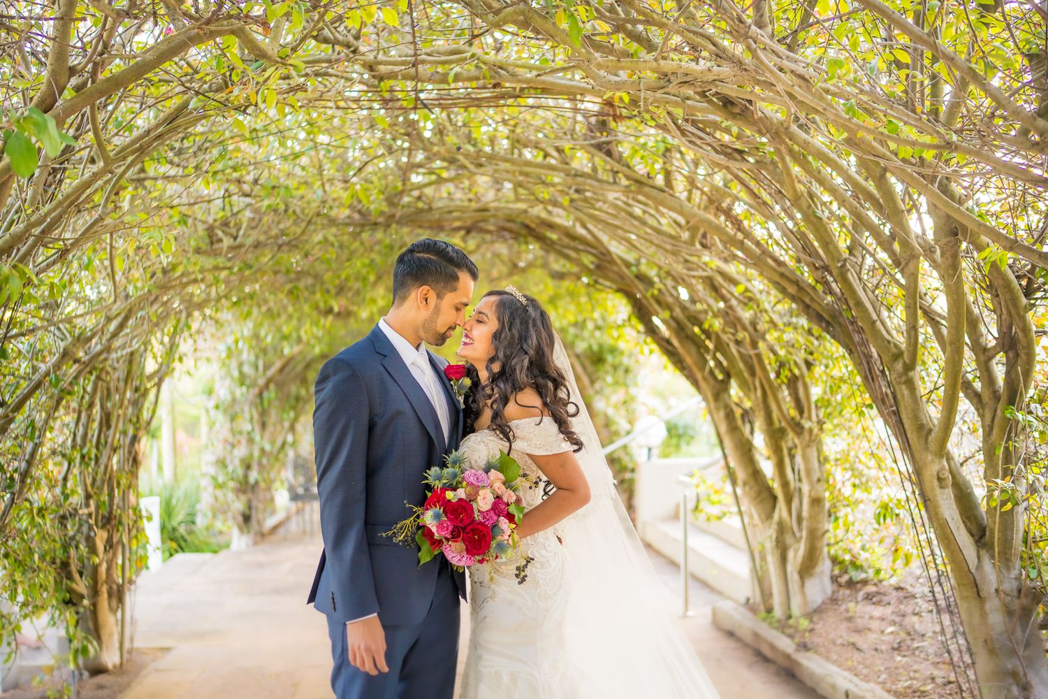 A romantic wedding photo under a natural archway of trees with a bride holding a red bouquet.
