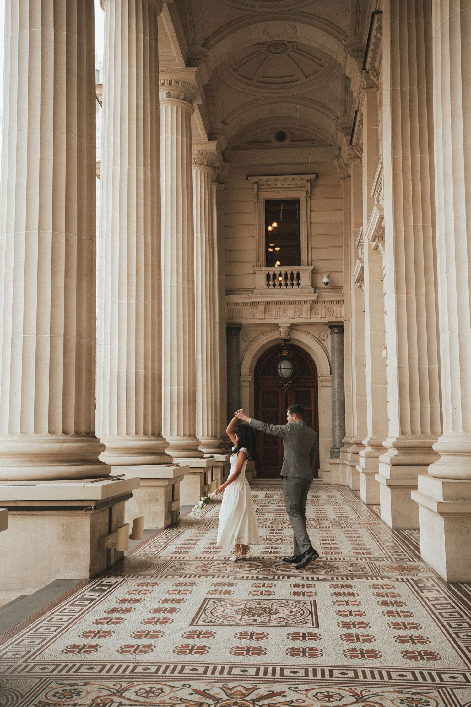 Bride and groom dancing among grand classical columns on ornate mosaic tile floor in elegant historic building.