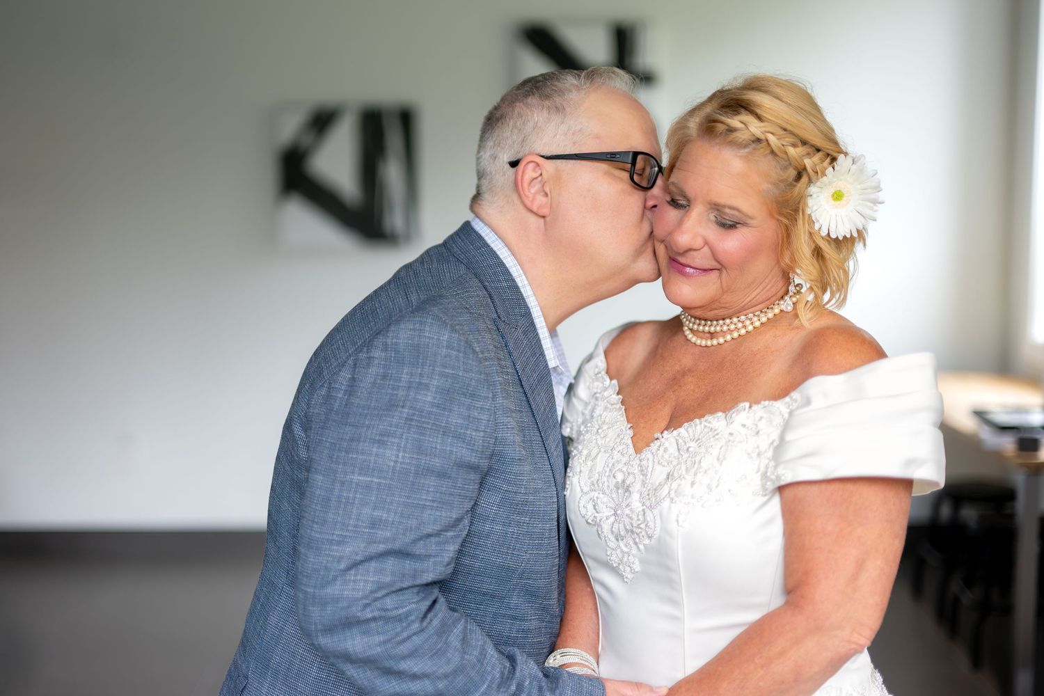 Groom in gray suit kissing bride in white off-shoulder dress with pearl necklace and flower in hair.