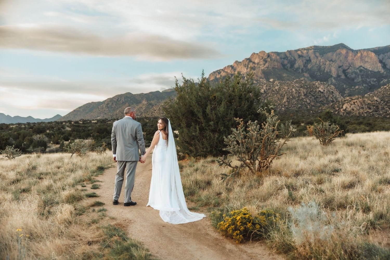Bride and groom walk a desert path with Sandia Mountains backdrop in golden hour light.