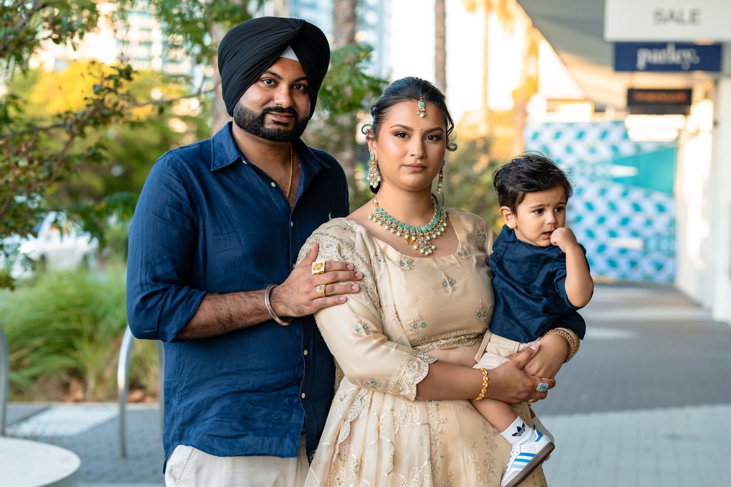 South Asian family in traditional attire, man in turban, woman in embroidered lehenga, holding a toddler outdoors.