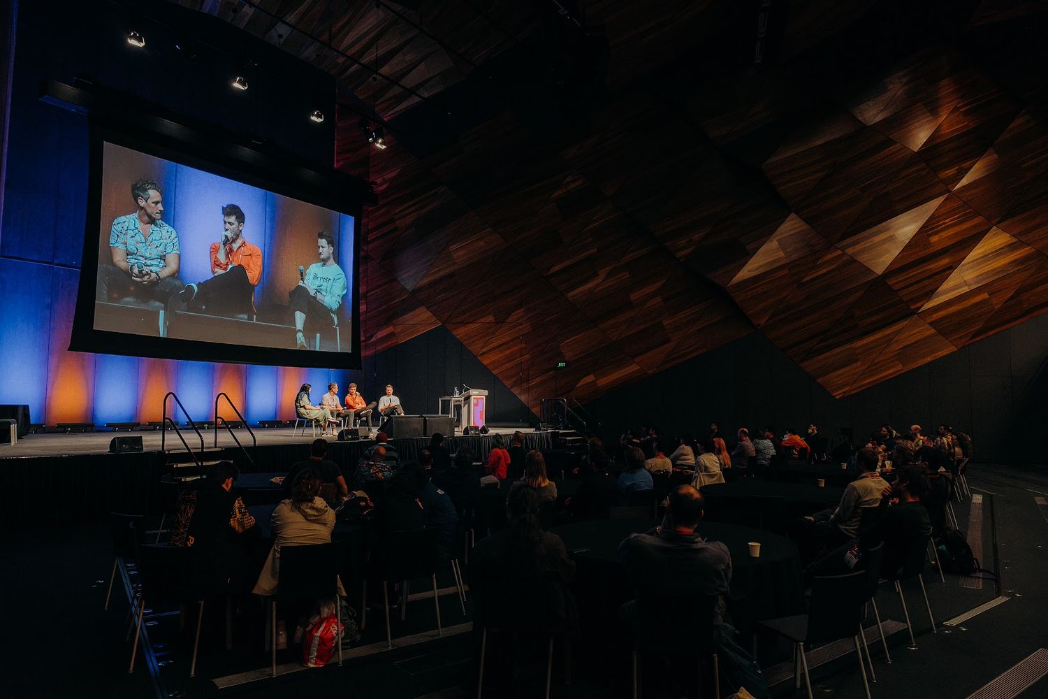 A large conference audience watches a panel discussion on stage with a big screen displaying speakers at a modern venue.