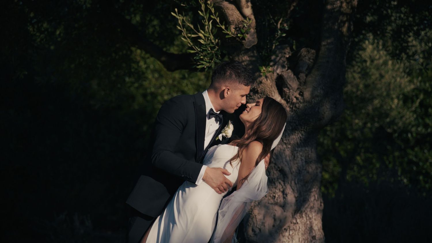 A bride and groom share a romantic kiss beside a large tree in a lush, dimly lit outdoor setting.