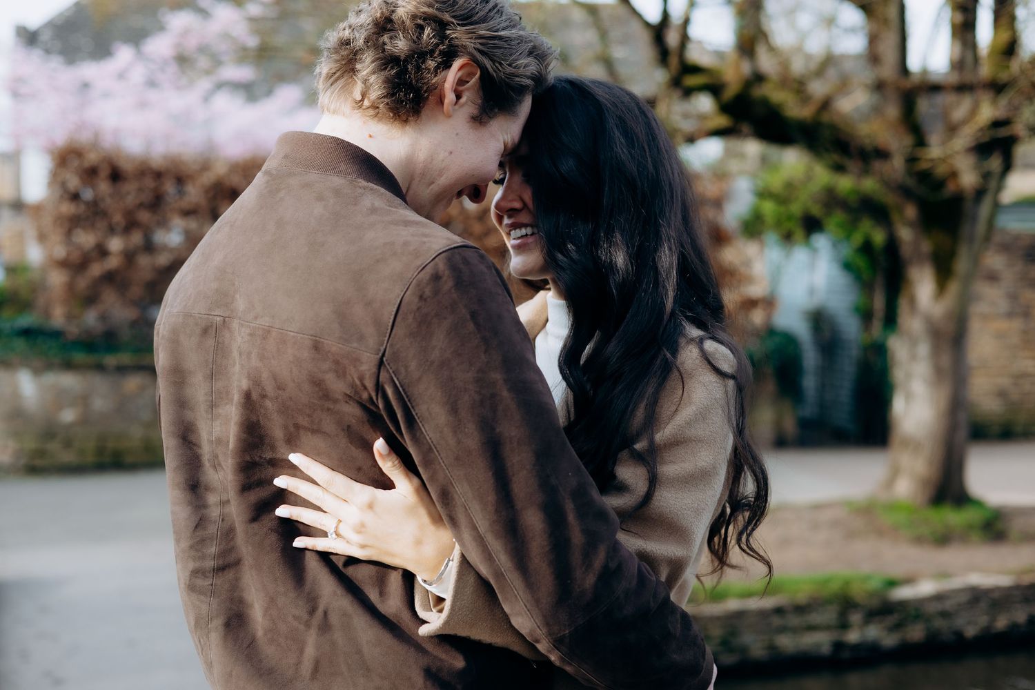 A happy couple embraces and smiles outdoors near bourton on the water for their cotswolds engagement photoshoot.