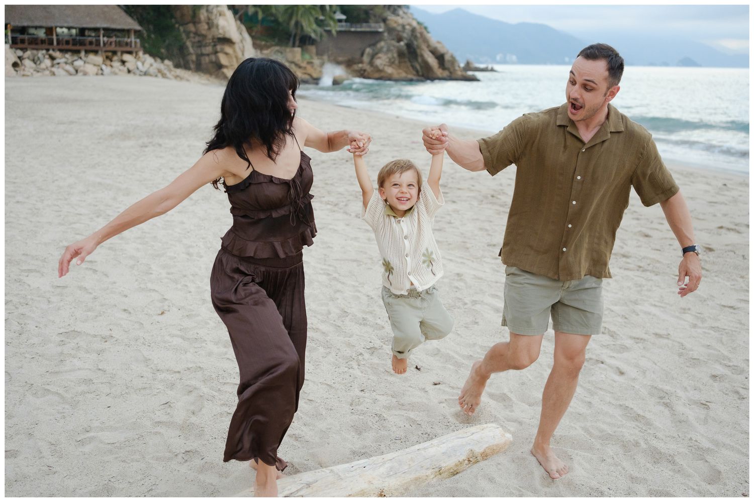 A family plays together on a beach, lifting a child in the air while wearing white clothing in black and white photography.