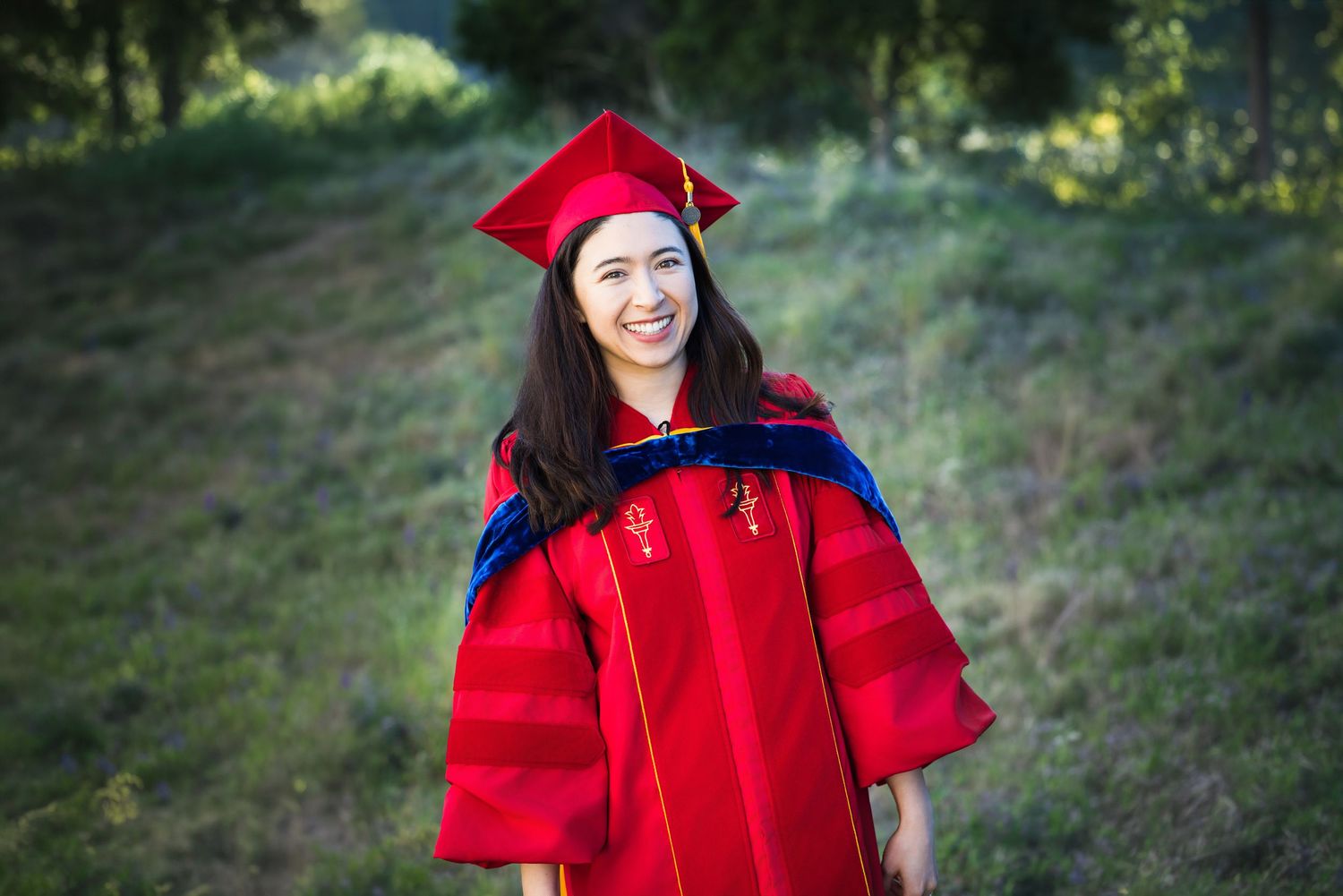 Smiling young woman in red graduation cap and gown standing outdoors on a green hillside.