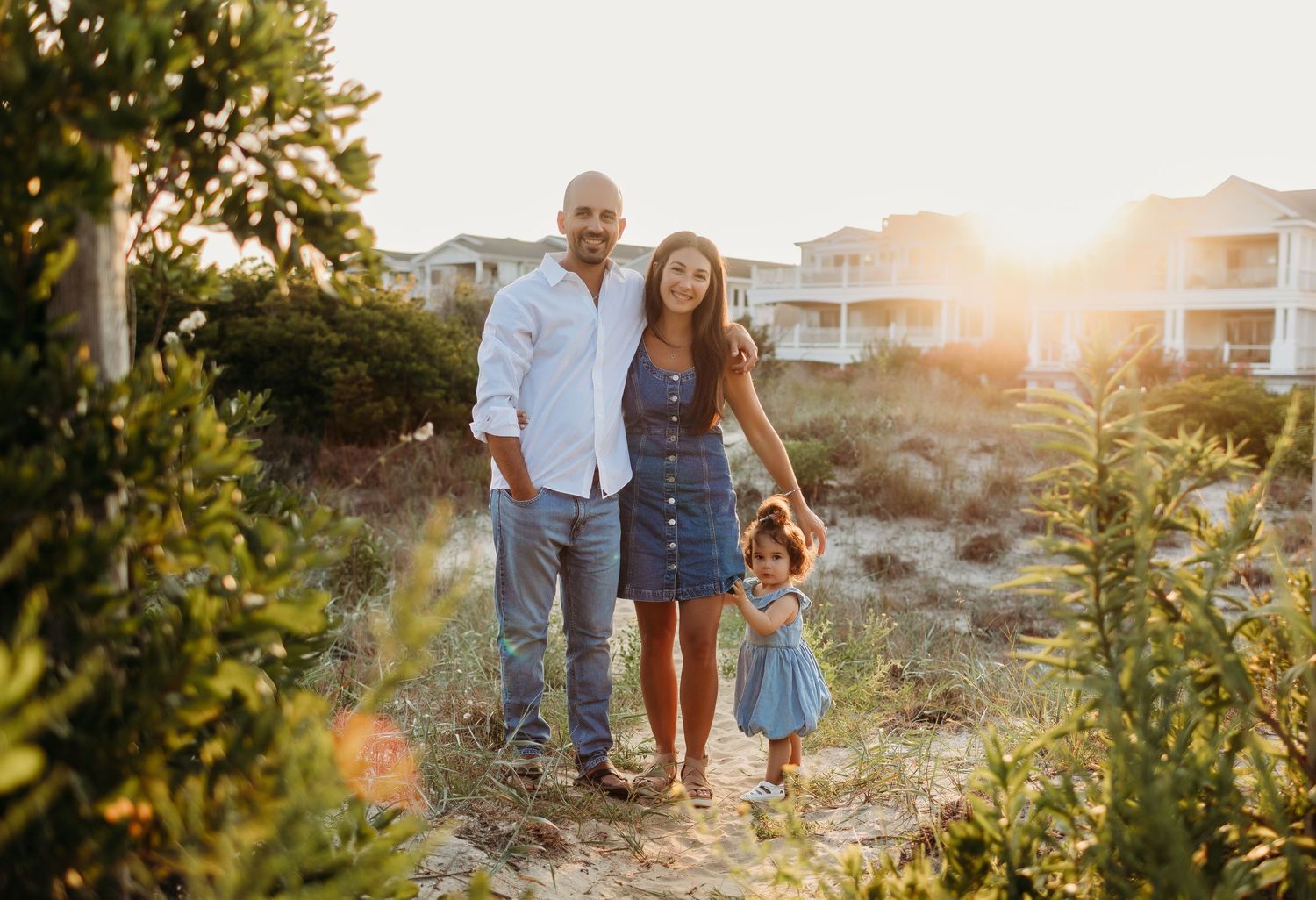 Family of three posing at golden hour on a sandy beach path surrounded by lush greenery and coastal homes.